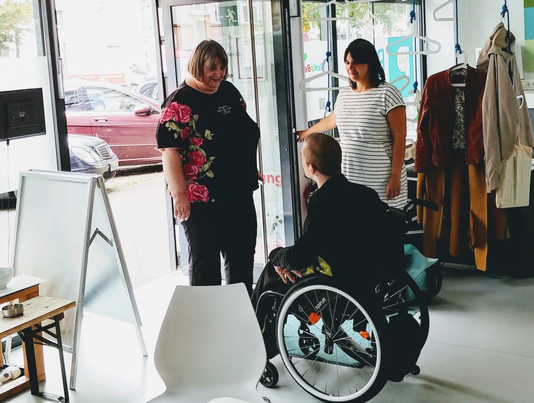 Three people - two women and a man in a wheelchair - are standing in the PIKSL entrance area. The man greets the woman entering through the door. 