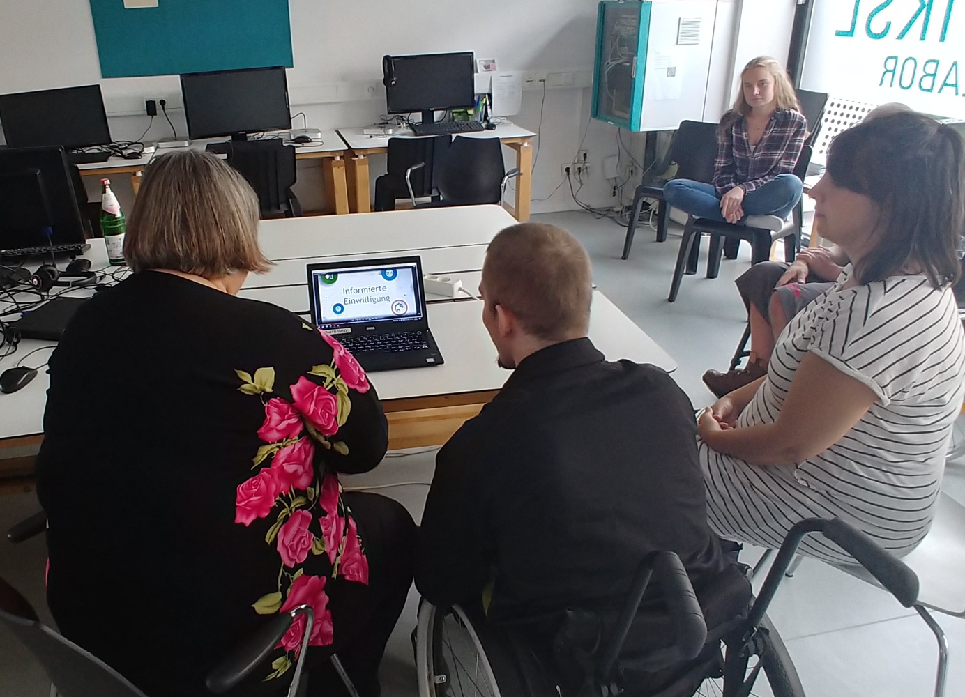 Monika and Dominik sit at a table in front of a laptop. A video of the Informed Consent is opened on the laptop.