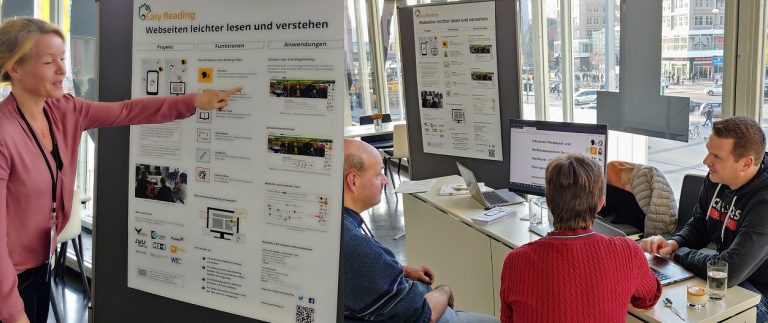 Frau zeigt auf ein Plakat, Mann sitzt mit zwei Besuchern am Messestand und zeigt das Easy Reading Programm