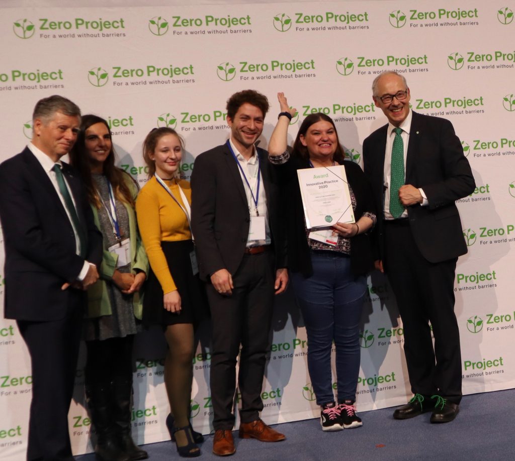 Three men and three women stand side by side and smile into the camera. One of the women is holding the award certificate in her hand. In the background is a wall with Zero Project logos.