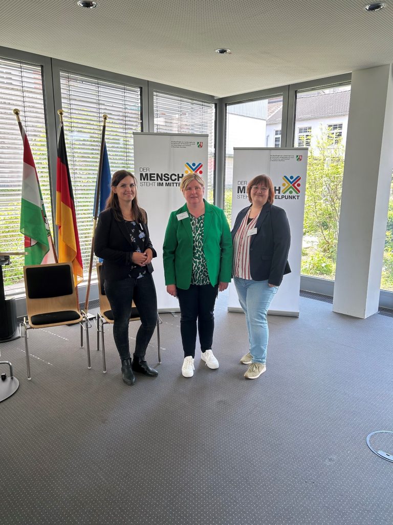 Drei Frauen stehen in einem Raum mit zwei Fensterfronten vor zwei Roll-Ups. Auf den Roll-Ups steht "Der Mensch steht in der Mitte". Links neben den Roll-Ups stehen drei Flaggen. Die Flagge von NRW, die Flagge vn Deutschland und die Flagge von Europa. 