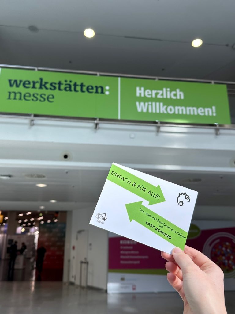 Im Vordergrund: Hand die eine Postkarte hochhäldt auf der steht: Einfach und für alle! Das Internet barrierefrei erleben mit Easy Reading 
Im Hintergrund: Eine Messehalle mit einem Eingang. Über dem Eingang hängt ein Banner auf dem steht Werkstätten:Messe Herzlich Willkommen! 