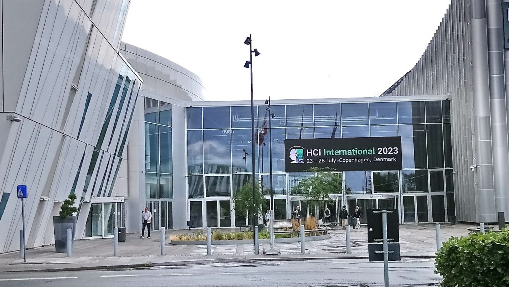 Konferenzzentrum dre HCI in Kopenhagen. EIn Gebäude mit gläserner Front an dem ein Banner mit der Beschriftung HCI International 2023 23. bis 28. July, Copenhagen, Denmark steht. 